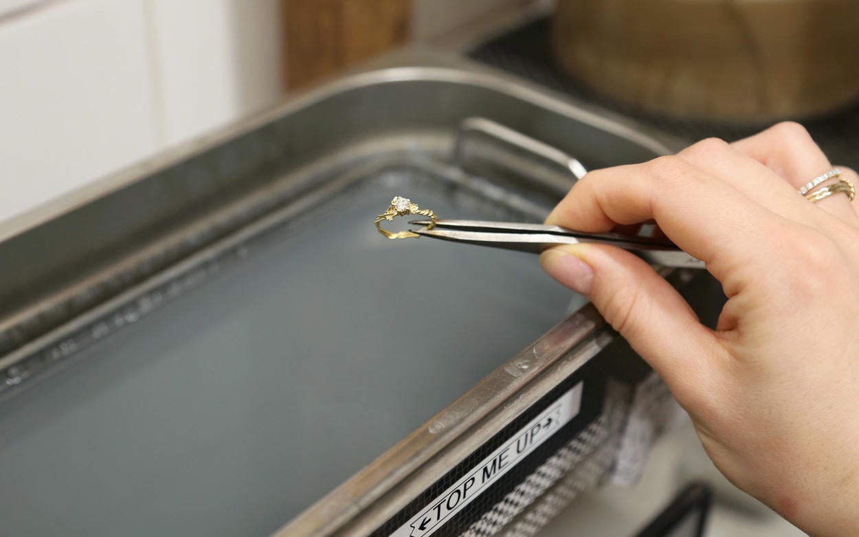 A gold ring held in jewellery tweezers held over the water bath of an ultrasonic cleaning machine.