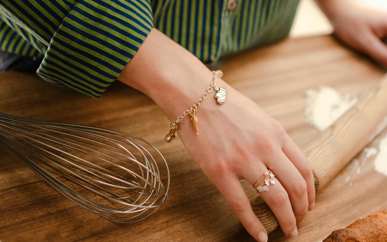 A woman wearing Alex Monroe baking charm bracelet using a rolling pin to roll dough on a wooden work top.