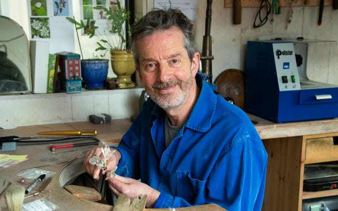 Alex Monroe at his workbench wearing his blue jewellers' jacket, holding two of his small silver floral stems.