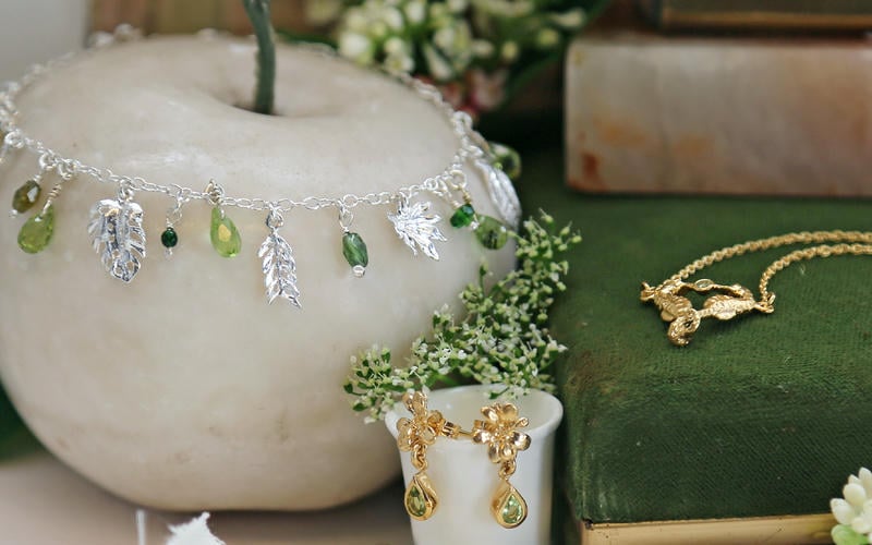 Peridot and silver leaf charm bracelet displayed on a white porcelain apple, next to a pair of seahorses necklace.