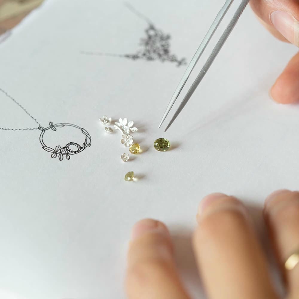 A Jeweller using tweezers to arrange loose yellow and green coloured gemstones and silver castings on a piece of paper, into the shape of design sketch for a necklace.