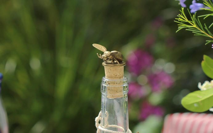 Bumblebee brass bottle stopper in a clear white glass bottle with green foliage and pink and purple flowers.