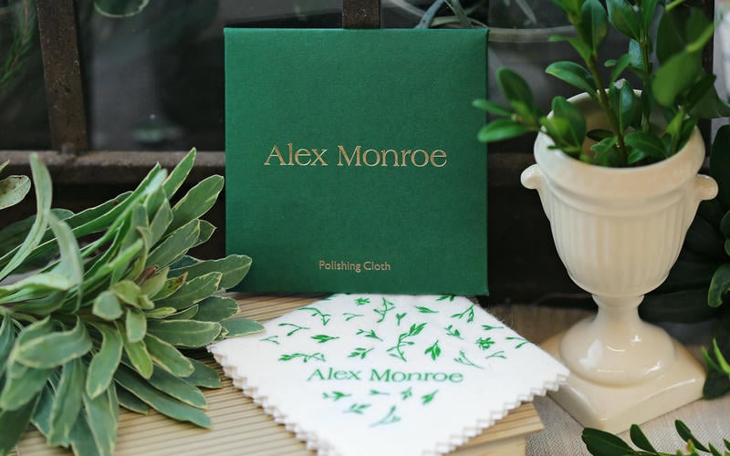 Alex Monroe branded jewellery silver polishing cloth displayed on a table amongst green plants.