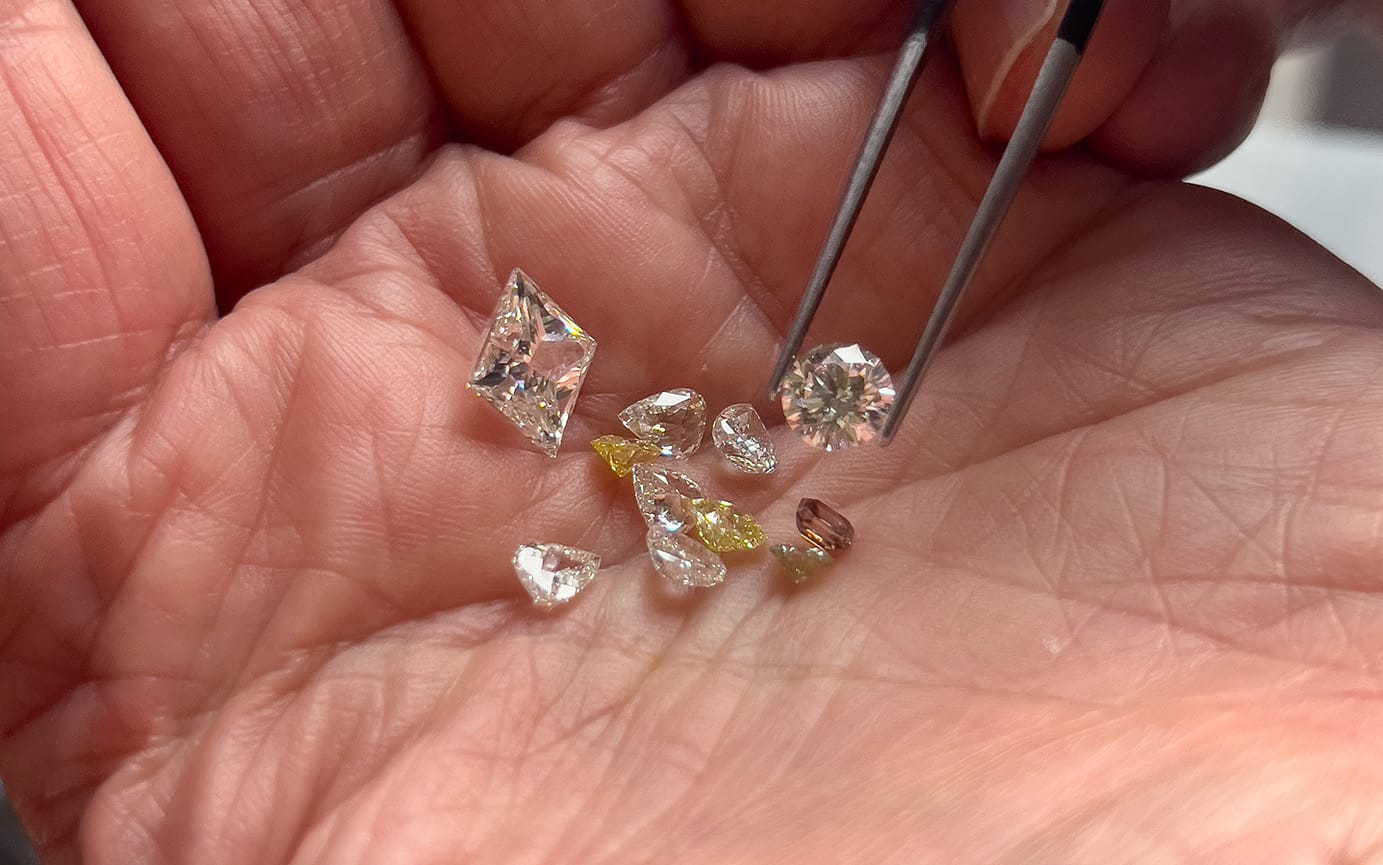 A selection of different white, yellow and champagne coloured diamonds held in the palm of a hand, with one being picked up with tweezers.