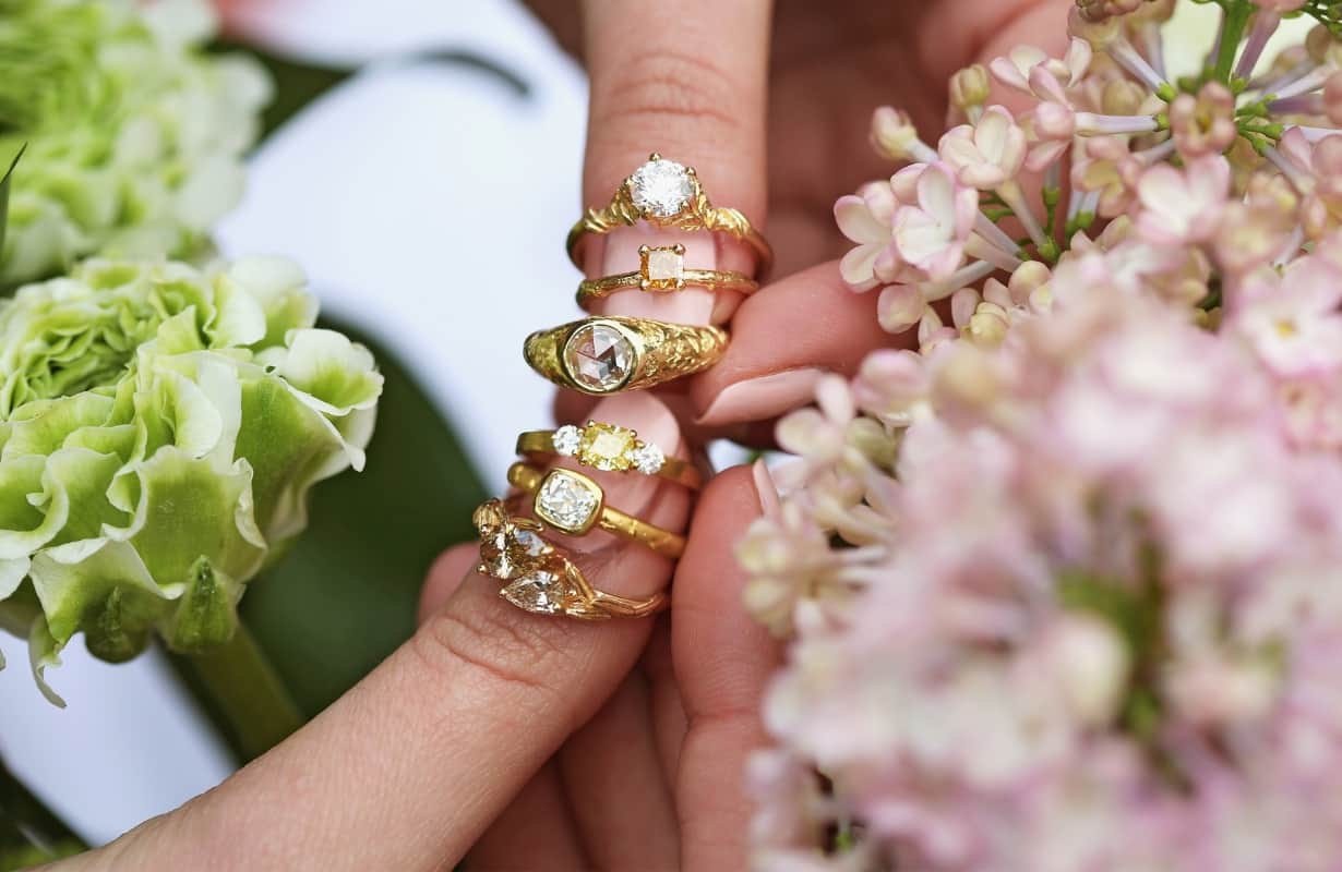 Assortment of six diamond rings, held across two thumbs. Some are large solitaire diamonds, others coloured diamonds, with smaller white diamonds.
