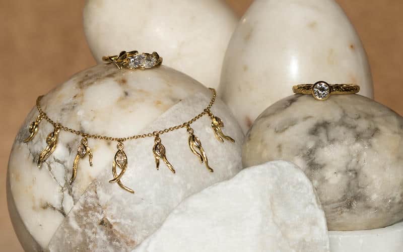Statement solid gold diamond charm necklace, and solitaire diamond ring, displayed on marble spheres.