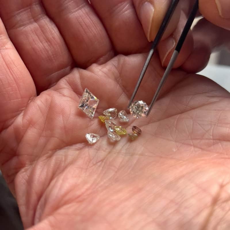 A selection of white and champagne diamonds cupped in the palm of a hand, being inspected with tweezers.