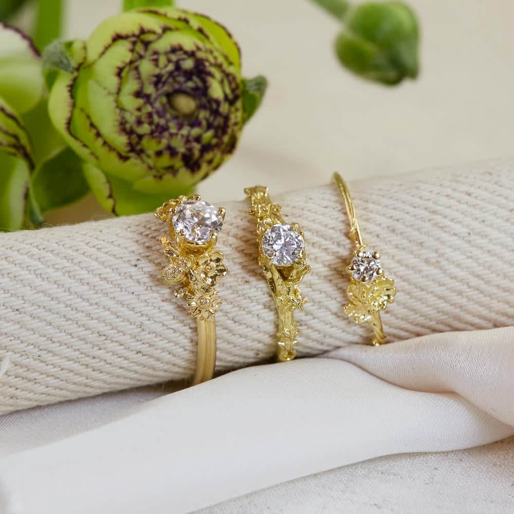 Three gold engagement rings with intricate floral detailing and sparkling central stones, displayed on a rolled cream napkin with a green flower in the background