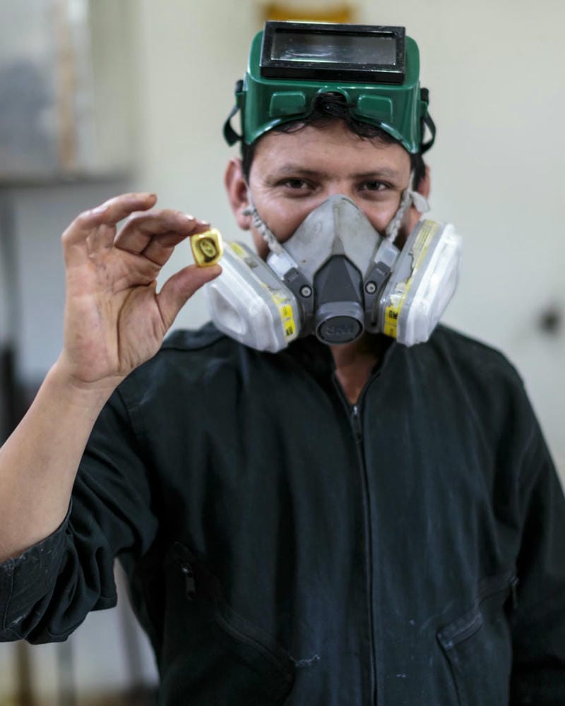 Miner in dark blue overalls wearing a respirator mask and safety visor propped on his head, holding a cube of processed fairmined gold.