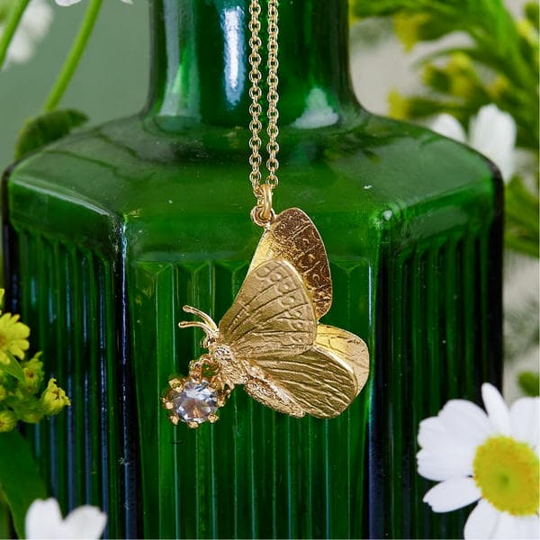 Butterfly necklace with amethyst gemstone in front of hexagonal green glass bottle.