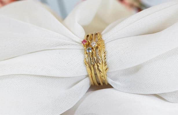 Four fine yellow gold rings, three with a single gemstone and one with a slim feather design, with a piece of white fabric pulled through the centre.