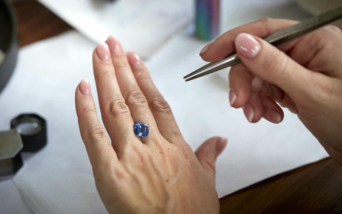 A large blue sapphire held on the back of a hand, resting between two fingers, with a pair of tweezers poised to pick it up.