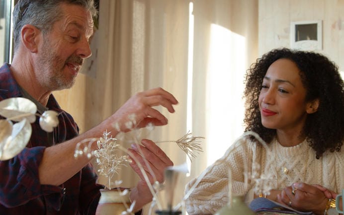 Silver floral stems arranged in a stoneware vase and a blue glass bottle on Alex's workbench in dappled sunlight.