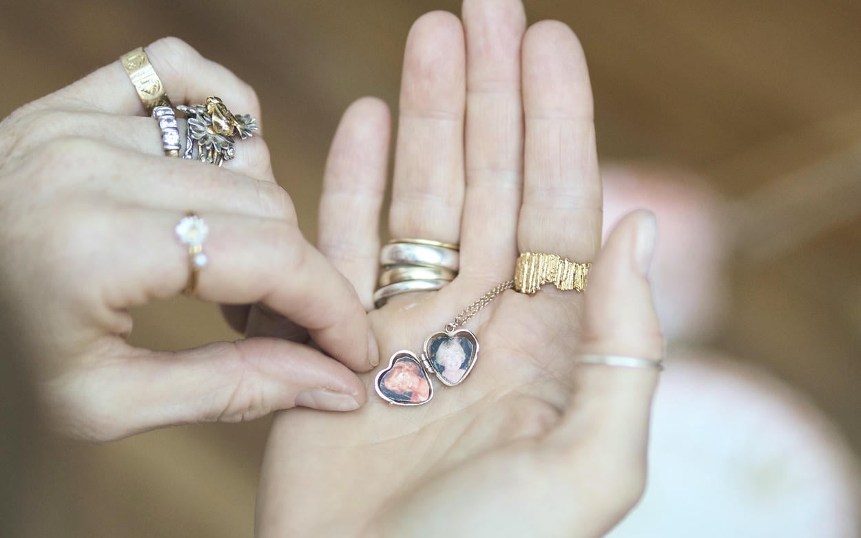 Womans open hands adorned with various silver and gold Alex Monroe rings, holding a small open gold heart shaped locket with photos inside.