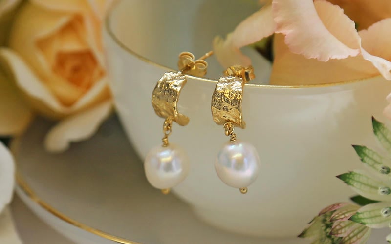 Pair of bark textured gold plated huggie hoop earrings with large irregular pearls balanced on the side of an ornate teacup.