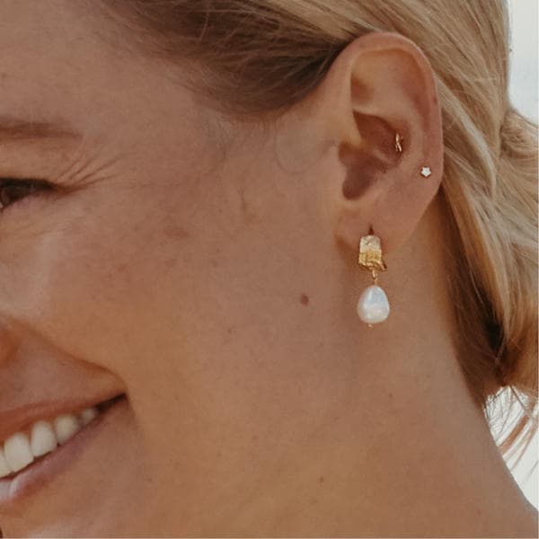 Woman wearing large irregular pearl huggie drop earrings.