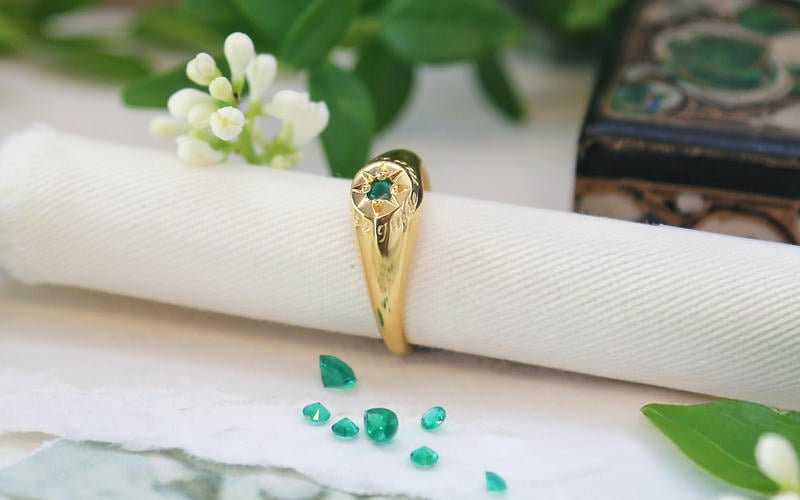 Emerald star-set signet ring on a roll of parchment paper with white flowers and green leaves surrounding.
