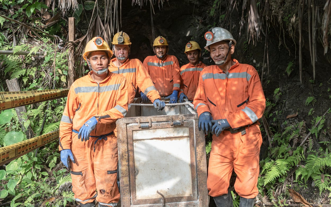 A group of miners in orange jump suits with hard hats posing near a mine entrance.