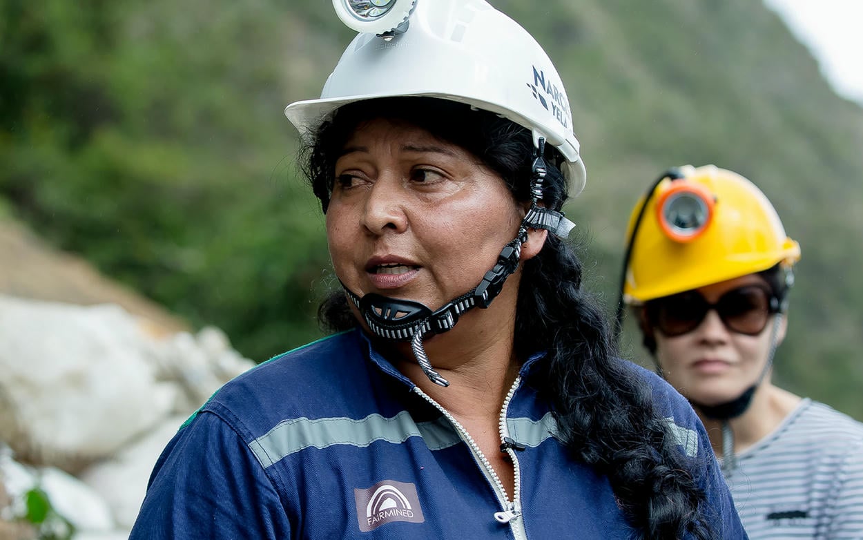 A woman wearing a blue Fairmined Gold logo'd miners jump suit with white hard hat and light.