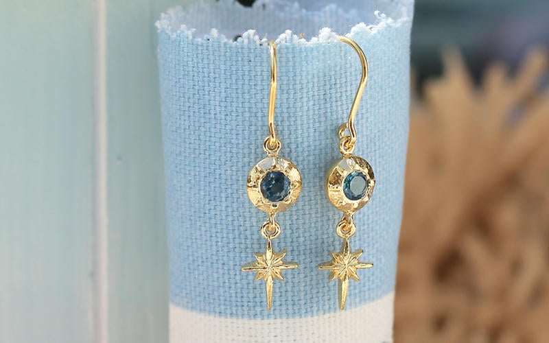 Blue topaz star drop earrings with pastel blue beach hut style wooden panels and blue fabric.