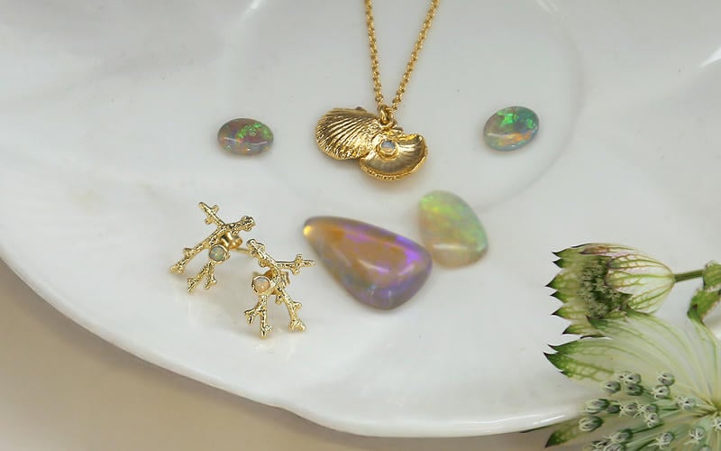 Scattered, loose rainbow pearlescent opal gemstones with a pair of solid gold opal coral texture stud earrings and a clamshell necklace.