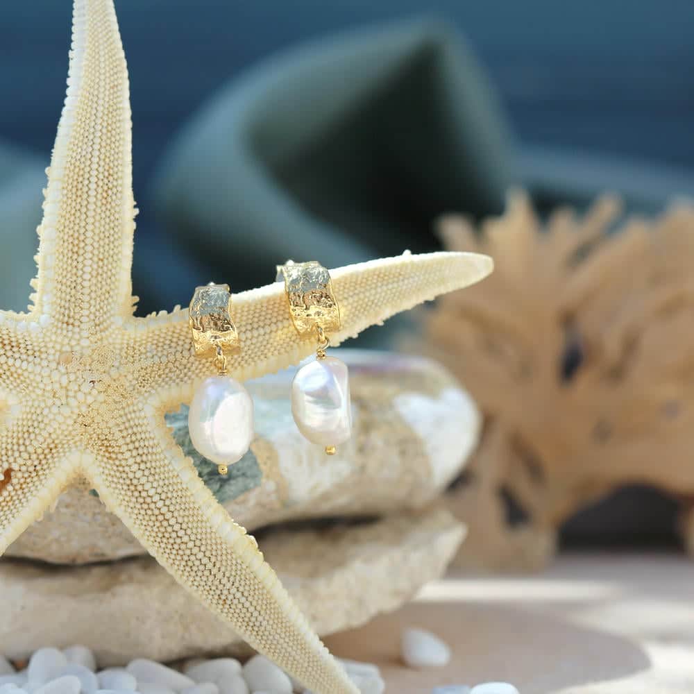 Gold and pearl drop earrings displayed on a starfish, with a coastal backdrop of shells and coral elements.