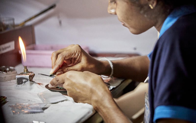 A close up on a womans hands as she works on adding facets to a dark green gemstone using hand tools and a flame
