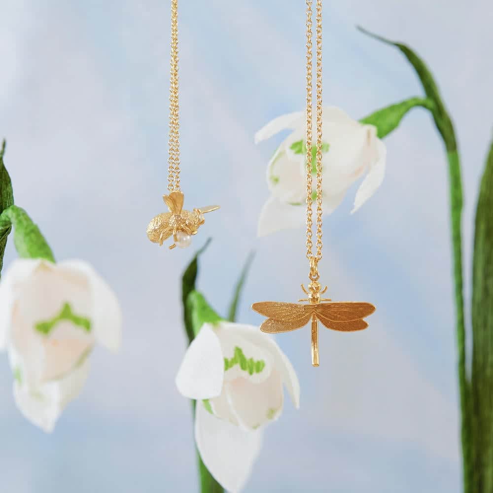 Gold bee and dragonfly necklaces hanging in front of delicate white snowdrop flowers, with a soft blue background