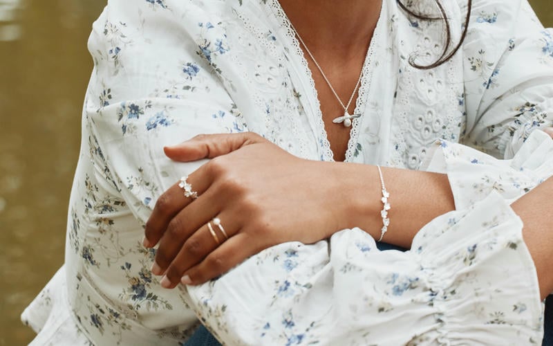 A woman wearing a white blouse with subtle blue flower print. She wears a silver baby bee necklace, forget me not bracelet and ring.