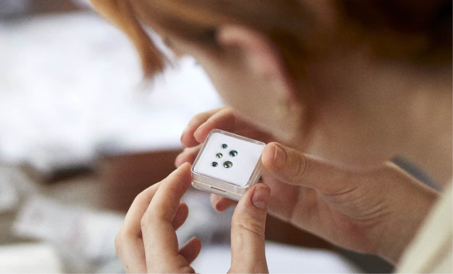 A woman inspecting a small clear container with 5 green blue sapphires cushioned inside.
