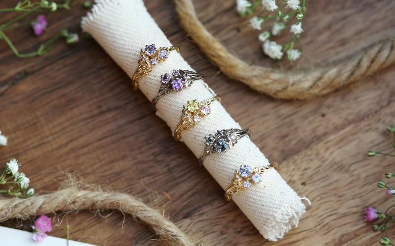 Selection of 5 trilogy solid yellow and white gold coloured gemstone rings on a roll of hessian with pink and white flowers forming a border.