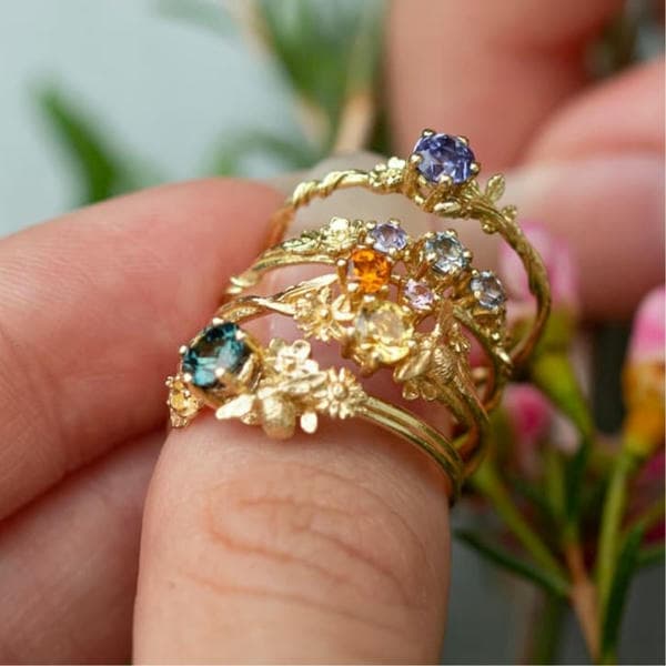 Stack of coloured sapphire solid gold rings being held in front of some pink flowers.