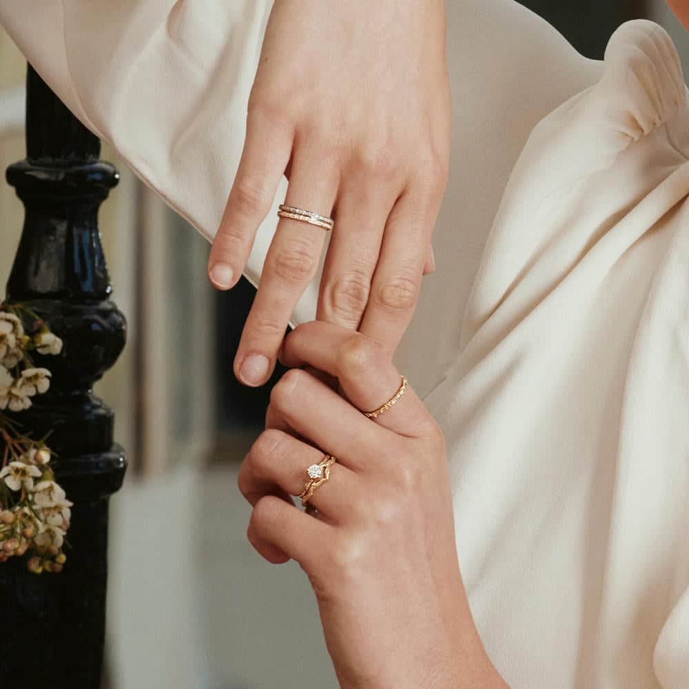Close-up of two hands wearing elegant gold and diamond rings, including wedding bands and a floral-inspired engagement ring, with a soft cream blouse in the background.