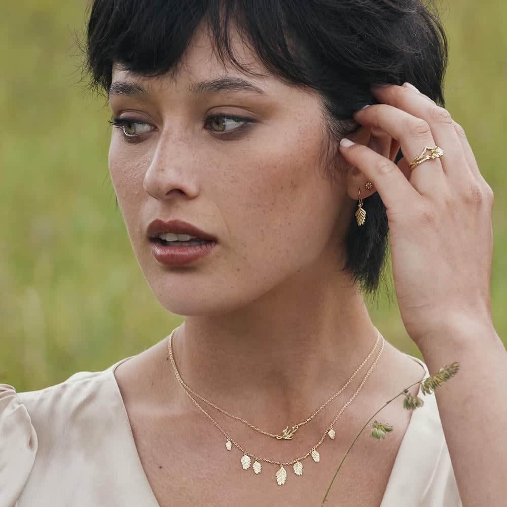 Model wearing layered gold necklaces with leaf and bird charms, a gold leaf earring, and matching rings, styled with a cream blouse in an outdoor meadow setting.