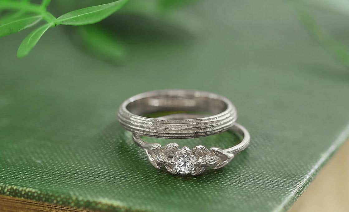 A white gold wide column textured wedding ring resting on a white gold grass seed desgin solitaire diamond engagement ring, on a textured green vintage book cover with leaves in the background.