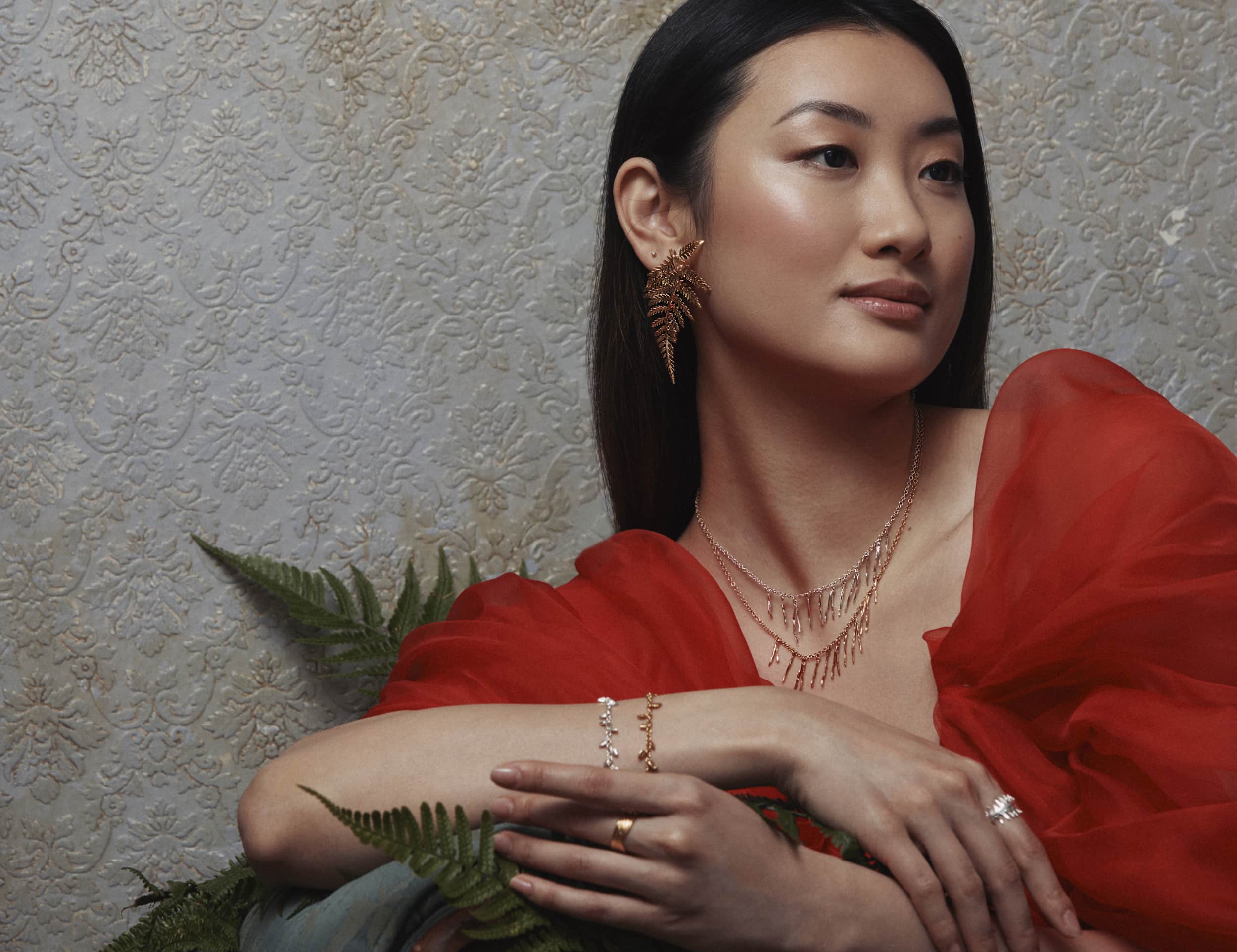 Woman in a red dress with billowing sleeves sat on a green sofa, wearing large fern leaf stud earrings.