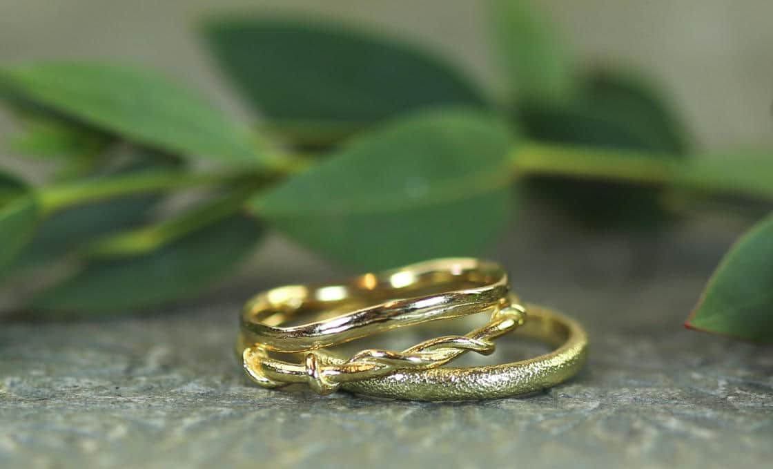 Close up of a stack of three different yellow gold wedding rings, a stippled texture band, wavy band and weaving vine style band, with green leaves in the background.